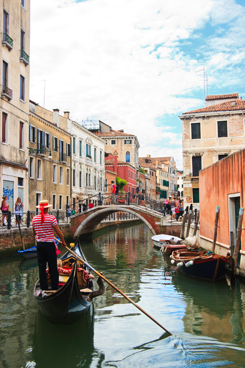 Venice, Italy - August 6th 2010: A gondolier rowing his gondola with passengers towards the bridge in Campiello del Cristo. Tourists sightseeing and taking photo along the canal.