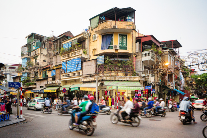 Busy street corner in old town Hanoi, Vietnam. Lots of people are commuting on motorbikes or cars. The street is lined by stores and appartment buildings.